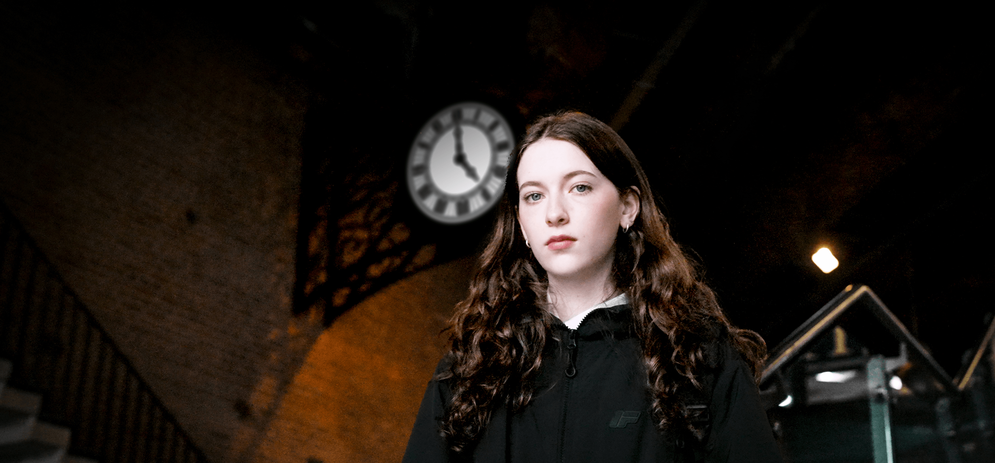A girl looking serious stood on the stairs of a train station below a clock on a brick wall.
