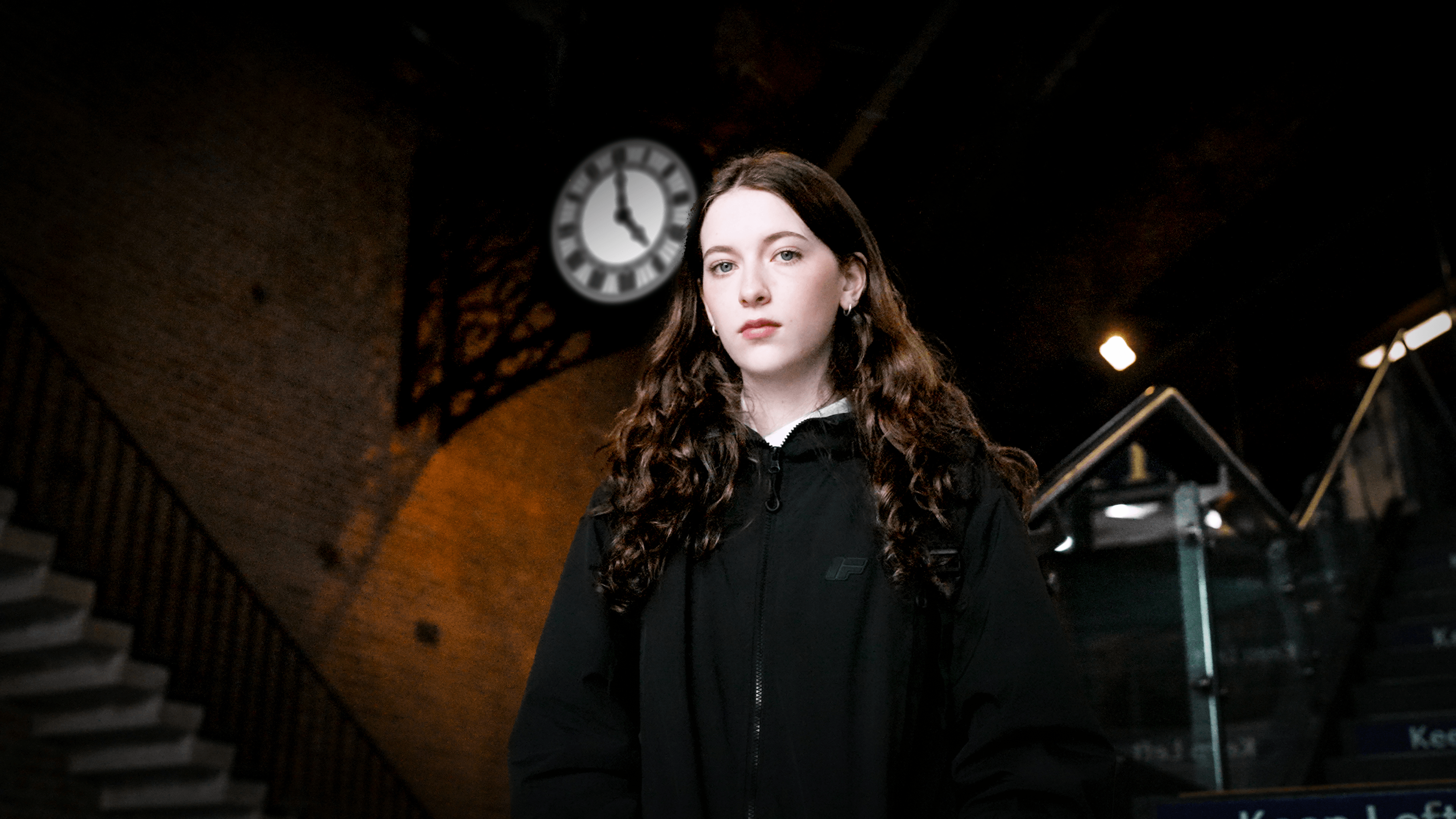 A girl looking serious stood on the stairs of a train station below a clock on a brick wall.