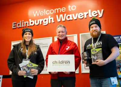 Railway Children members of staff wearing branded clothing stood either side of a member of staff from LNER in front of a red background which reads Welcome To Edinburgh Waverley.