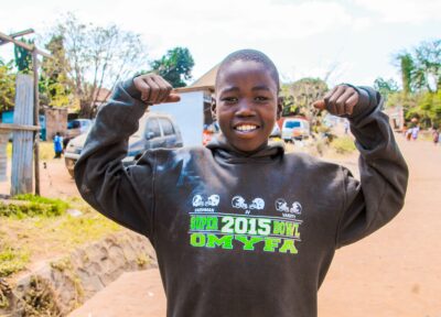A young boy in Tanzania wearing a dark grey hoodie with green and white writing raising his arms up and smiling.