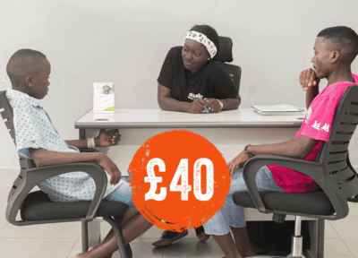Two young individuals sit across from a staff member at a child support desk, engaged in a supportive consultation or counselling session. The room has a clean, professional setting with chairs, notepads, and an informational desk calendar. The interaction reflects youth guidance, wellbeing support services, and community outreach programmes. At the forefront, a bright orange graphic with “£40” signifies the associated contribution level.