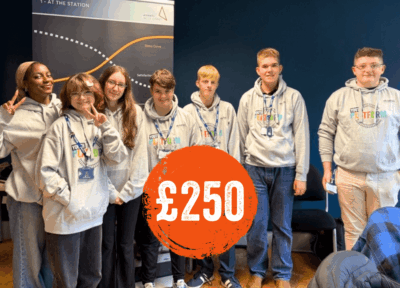A group of teenagers stand together wearing matching grey hoodies branded with a youth programme design. They appear to be in an indoor training or workshop environment, with banners and materials in the background. The group setting represents leadership development, youth empowerment, and educational training initiatives. A prominent orange circle featuring “£250” highlights the donation value linked to the programme.