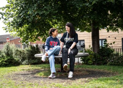 A Railway Children youth practitioner and a child sit together on a circular outdoor bench in a leafy park area, surrounded by green grass, shrubs and a large tree providing shade. Behind them, a residential brick building and metal railings are visible. The scene captures a calm outdoor moment in a community setting, highlighting nature, urban greenery and supportive interaction in a neighbourhood environment.
