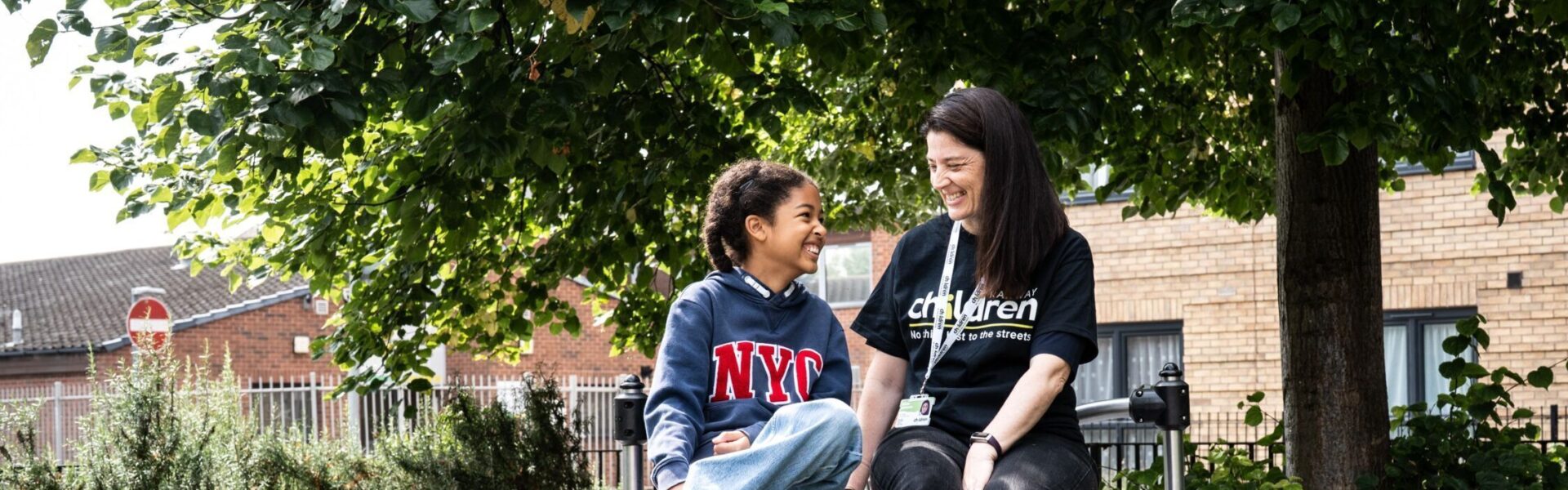 A Railway Children youth practitioner and a child sit together on a circular outdoor bench in a leafy park area, surrounded by green grass, shrubs and a large tree providing shade. Behind them, a residential brick building and metal railings are visible. The scene captures a calm outdoor moment in a community setting, highlighting nature, urban greenery and supportive interaction in a neighbourhood environment.
