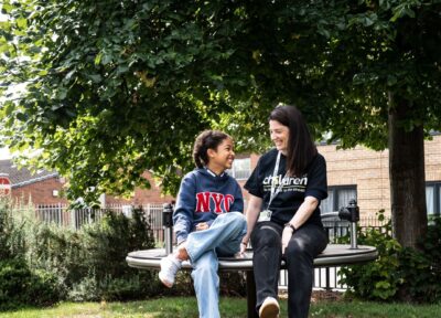 A Railway Children youth practitioner and a child sit together on a circular outdoor bench in a leafy park area, surrounded by green grass, shrubs and a large tree providing shade. Behind them, a residential brick building and metal railings are visible. The scene captures a calm outdoor moment in a community setting, highlighting nature, urban greenery and supportive interaction in a neighbourhood environment.