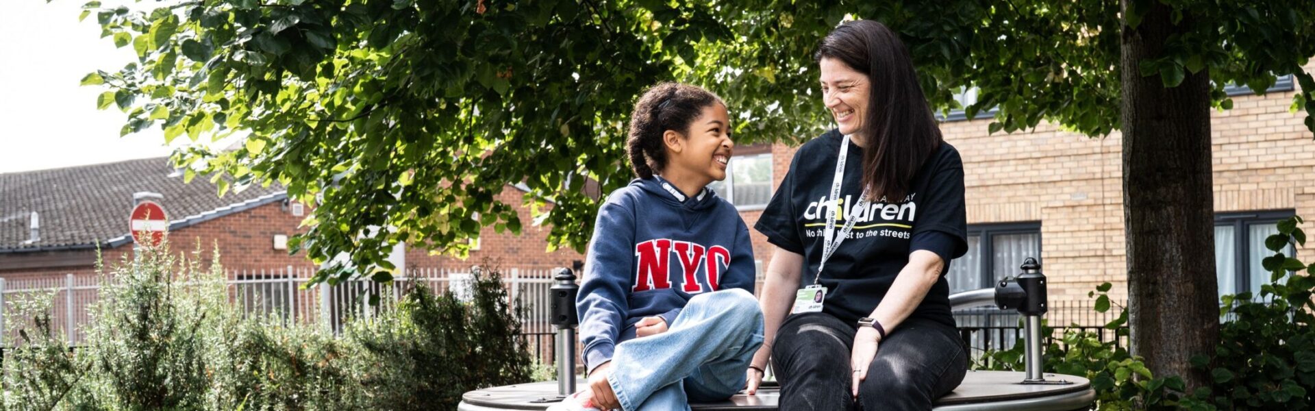A Railway Children youth practitioner and a child sit together on a circular outdoor bench in a leafy park area, surrounded by green grass, shrubs and a large tree providing shade. Behind them, a residential brick building and metal railings are visible. The scene captures a calm outdoor moment in a community setting, highlighting nature, urban greenery and supportive interaction in a neighbourhood environment.