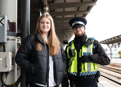 A teenage girl stood at a railway station platform in a black coat with a British Transport Police Officer.