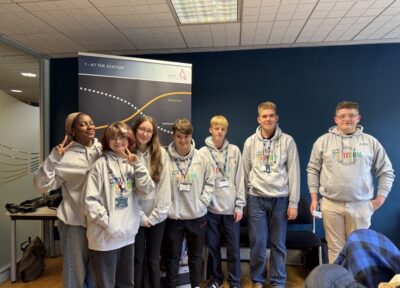 A group of students wearing matching grey Railway Children Youth Platform hoodies participate in an educational workshop, standing together in a modern office setting with informational signage in the background.
