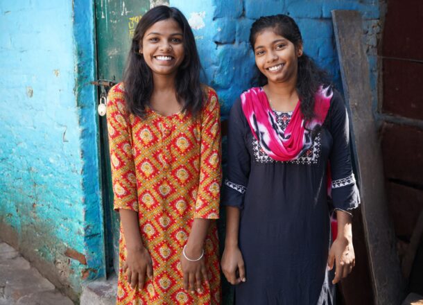 Two girls wearing brightly coloured clothes stood together smiling on the streets of India in front of a blue wall.