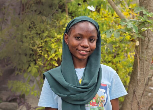A teenage girl in Tanzania wearing a white T-shirt and dark green headscarf, smiling at the camera in front of some trees and yellow flowers.