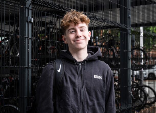 A teenage boy with red hair wearing all black and smiling in front of a fence and some bikes in the UK.