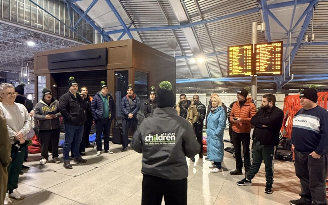 Large group of sleepout participants gathered in a circle inside a train station while listening to a briefing, with overhead information screens visible.