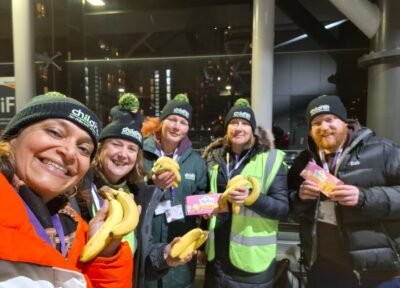 A group of five charity sleepout participants wearing matching black beanie hats and winter clothing standing together at night holding bunches of bananas and snack bars, with a city station structure in the background.