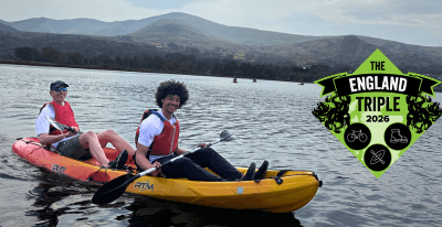 Two people in a kayak on a picturesque lake in the UK holding oars and smiling with an England Triple logo on the image.