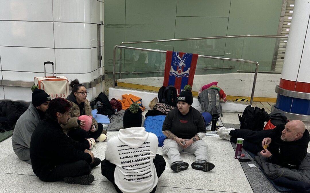 Small group sitting together on the floor indoors with sleeping bags and backpacks during a charity sleepout event, with a Crystal Palace flag hanging behind them.