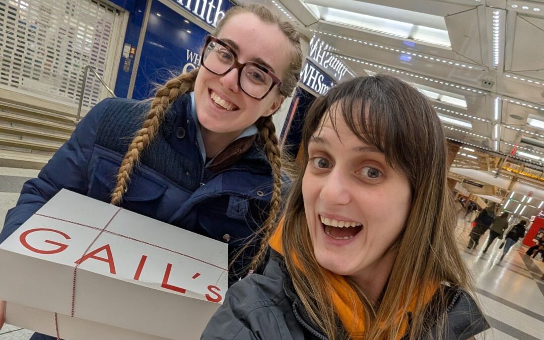 Two smiling participants holding large Gail’s Bakery boxes inside a brightly lit station concourse.