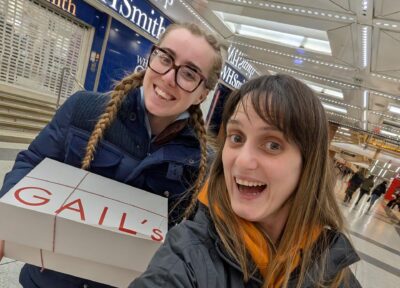 Two smiling participants holding large Gail’s Bakery boxes inside a brightly lit station concourse.