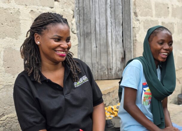 A Railway Children project worker wearing a black branded polo shirt sits beside a young person outside a rustic stone building. The setting features a weathered wooden door and items scattered on the ground, suggesting a rural or community environment. The image reflects grassroots outreach work, child support initiatives, and field‑based safeguarding programs delivered by Railway Children in vulnerable communities.