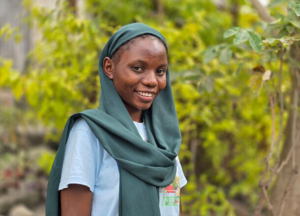 A girl stands outdoors in a natural, green environment, wearing a light blue T‑shirt and a deep green scarf draped over her head and shoulders. Behind her is lush foliage and soft natural light, creating a calm and rural backdrop. The scene reflects themes of community support, youth outreach, and humanitarian fieldwork, often associated with child‑focused development projects.