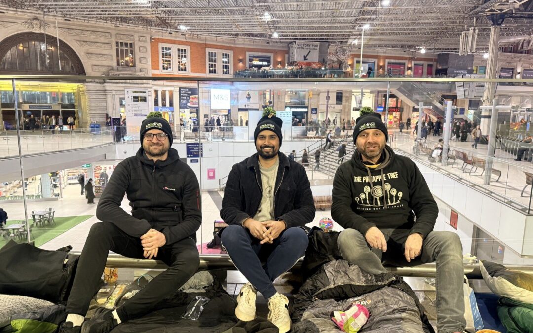 Three participants sitting on a raised indoor platform with sleeping bags and snacks, overlooking the busy concourse of a large train station.