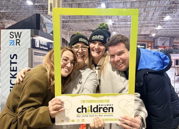 A group of four supporters standing together at London Waterloo station holding a green fundraising frame for Railway Children and a sign reading ‘Sleepout London Waterloo’. The group is dressed warmly for the event, with two individuals wearing Railway Children branded hats. Station signage and the concourse are visible in the background.