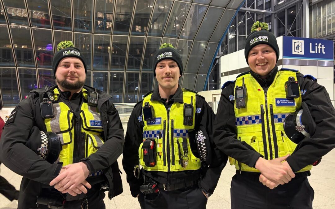 Three uniformed transport police officers wearing high‑visibility vests and charity-branded hats posing inside a modern station.