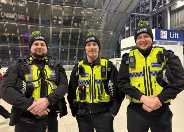 Three uniformed transport police officers wearing high‑visibility vests and charity-branded hats posing inside a modern station.