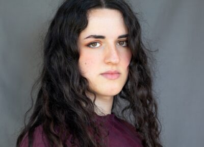 A young transgender woman in front of a grey background with dark hair and wearing a purple jumper.