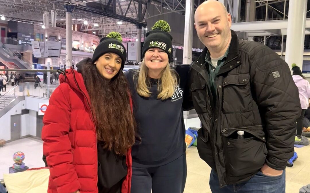 Three participants standing together indoors with sleeping mats and bags behind them inside a large, modern railway station.
