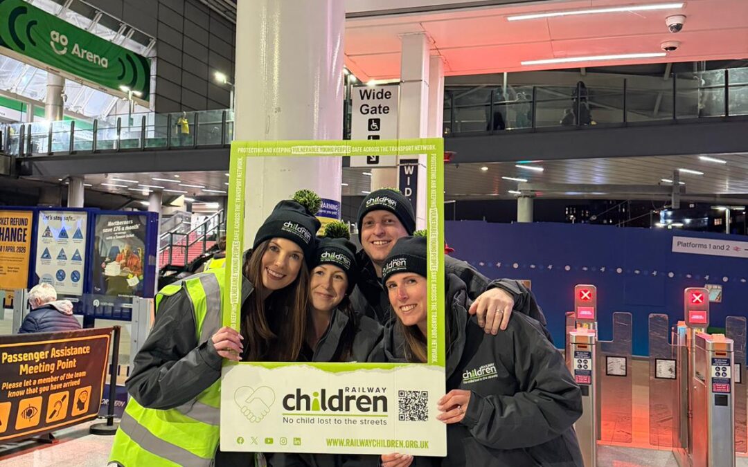 Four Railway Children staff members wearing charity-branded hats posing together and holding a large photo frame beneath a Manchester Victoria station sign.