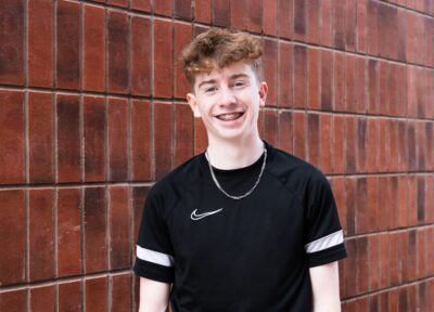 A young person stands against a textured red‑brick wall, wearing a black athletic T‑shirt with a white Nike logo and a thin silver chain necklace. The outdoor setting features evenly patterned brickwork that creates a clean, urban backdrop.
