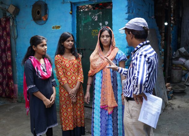 A Railway Children project worker stands in conversation with three individuals outside a blue-painted brick building in a narrow courtyard. Two teenage girls stand together on one side, while the mother stands between them and the project worker, dressed in patterned traditional clothing. The setting includes textured walls, a metal door, household items and an informal passageway, reflecting a low‑income neighbourhood where Railway Children’s frontline teams provide vital safeguarding and family‑support work. The scene highlights community engagement, protection of vulnerable young people, and collaboration with families in at‑risk urban areas.