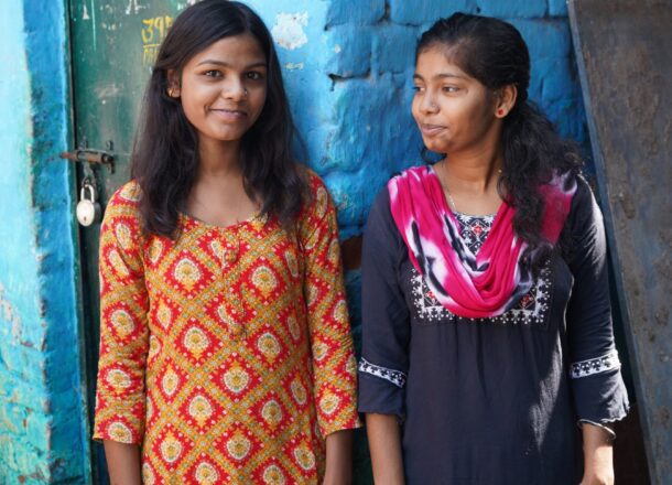 Two individuals stand outside a blue-painted brick building, wearing brightly patterned traditional clothing. The textured blue wall, metal doorway and surrounding alleyway create a vivid, urban setting often associated with community outreach and child‑protection work in low‑income neighbourhoods. The image highlights themes of resilience, local support programmes, and the environments where charities work to keep vulnerable children safe.