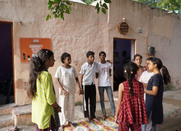 A group of young people are gathered outside a Railway Children community activity centre in India. They are standing together in a small courtyard area in front of a light‑coloured building, with one of the centre’s signs visible on the wall. Some of the young people appear to be talking or interacting as they stand in a loose circle on a patterned mat placed on the ground. Trees overhead cast shade across the scene.