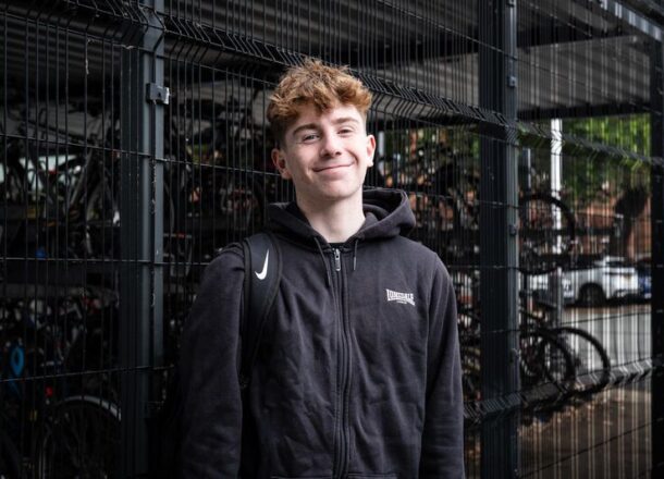 teenage boy in black hoody, standing outside a bike shed at a train station, smiling at camera