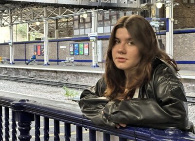 A child is leaning against a dark blue railing on a quiet railway station platform. The child is wearing a dark, oversized jacket with both arms resting on the railing. Behind the child, several railway tracks run through the station, bordered by platforms with seating areas and a row of colorful posters displayed on the opposite wall. The station features tall metal beams, overhead lighting, and a partially open roof structure that lets in natural daylight. A person is seated on a bench in the distance, adding to the calm, spacious atmosphere of the scene.