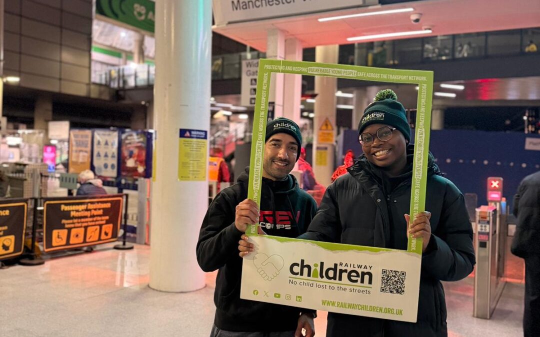 This photo shows two individuals standing inside Manchester Victoria train station, holding a large green promotional frame for “Railway Children,” a UK charity supporting vulnerable young people. The individuals are dressed in winter clothing, including hats branded with “Railway Children,” and stand near the station concourse beneath a prominent red and white Manchester Victoria signage board. The background includes ticket barriers, digital advertising screens, and station signage, creating a busy and well‑lit transport hub setting. The promotional frame features the Railway Children logo, website URL, and a visible QR code.