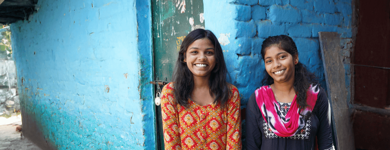 Two individuals stand outside a blue-painted brick building, wearing brightly patterned traditional clothing. The textured blue wall, metal doorway and surrounding alleyway create a vivid, urban setting often associated with community outreach and child‑protection work in low‑income neighbourhoods. The image highlights themes of resilience, local support programmes, and the environments where charities work to keep vulnerable children safe.
