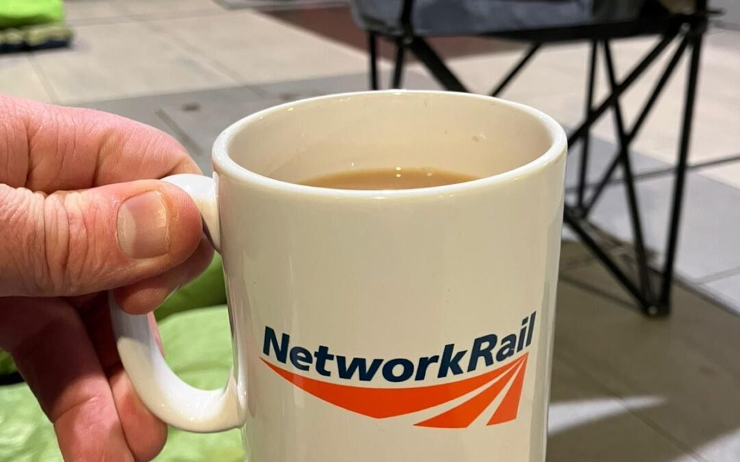 Close-up of a hand holding a Network Rail-branded mug filled with a hot drink, with sleeping bags and chairs in the background.