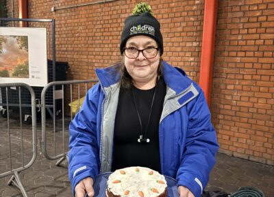 Person holding a homemade cake decorated with small carrots on top while wearing a charity beanie hat at an outdoor platform area.
