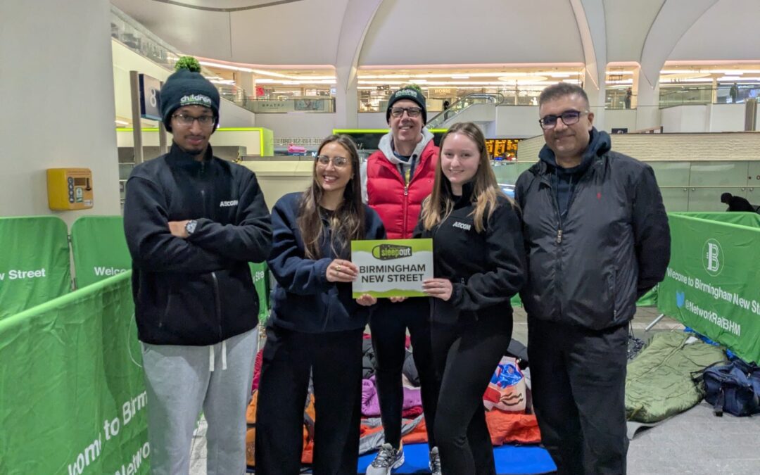 Group of five standing together at Birmingham New Street Station holding a Sleepout Birmingham New Street sign with colourful sleeping bags behind them.