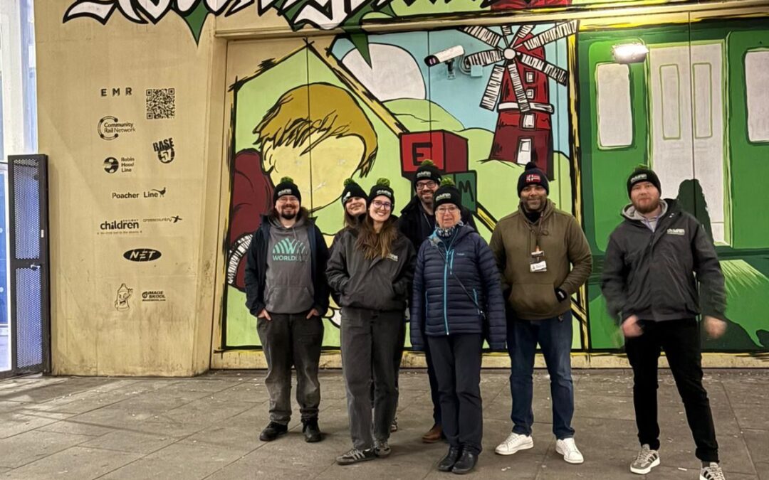Group of six people posing in front of a colourful mural reading “We Are Nottingham” at an outdoor location near a station.