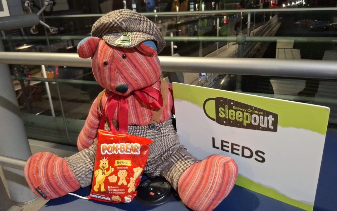 A patterned teddy bear dressed in vintage-style clothing placed beside a Sleepout Leeds sign with a packet of Pom-Bear snacks in front.