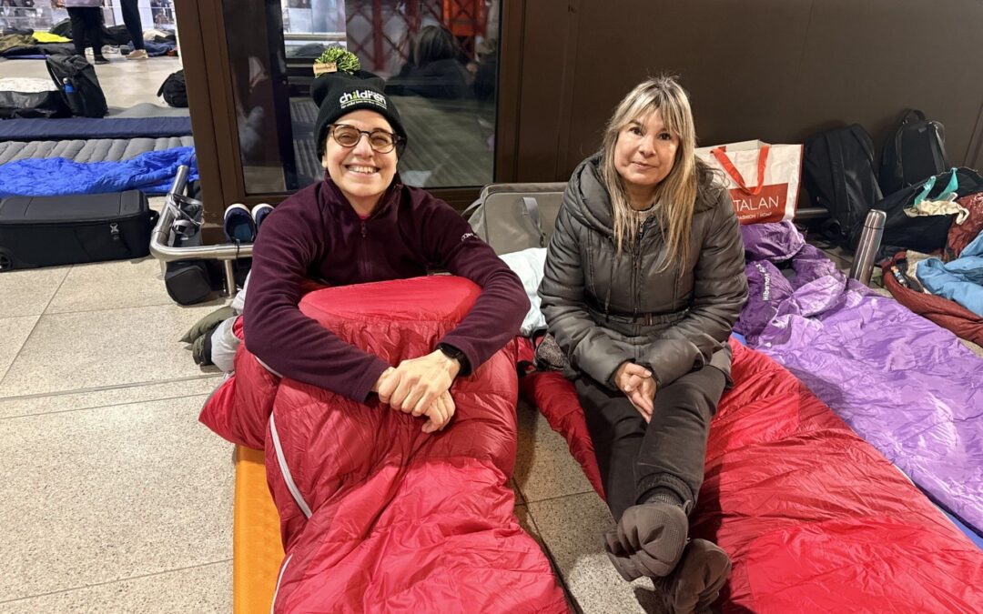 Two people sitting on the floor in sleeping bags at an indoor station area, surrounded by bags and sleepout gear.