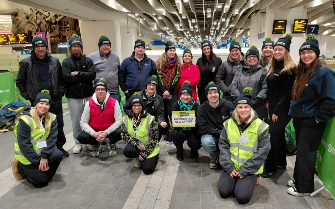 Large charity sleepout team wearing matching hats gathered inside Birmingham New Street Station, some kneeling at the front for a group photo.