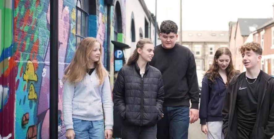 A group of people walk together down a narrow urban street lined with brick buildings and colorful street art. On the left side, a vibrant graffiti mural covers the wall, featuring bright abstract shapes and bold colors such as green, yellow, pink, and purple. The group appears to be engaged in conversation while moving along the sidewalk. They are wearing casual outdoor clothing including jackets, hoodies, and jeans. In the background, rows of terraced houses and a partially visible building create a typical city‑neighborhood atmosphere. The overall scene conveys community, youth engagement, and an urban creative environment.
