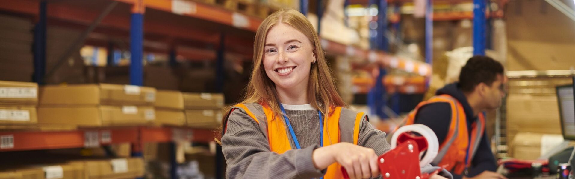 Girl with auburn hair in high visibility jacket in a warehouse wrapping boxes and smiling at the camera.