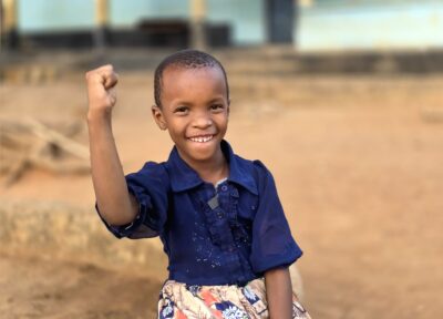 Young girl sitting outdoors on sandy ground, raising one arm with a closed hand, wearing a dark shirt and patterned skirt.