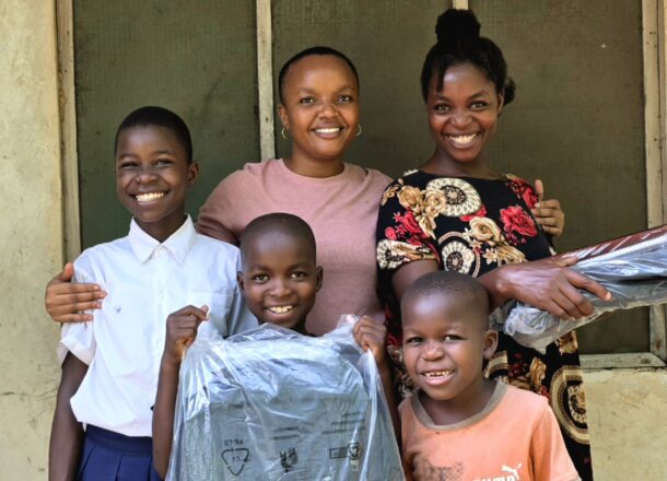 A group of smiling children stand with two adults outside a building, celebrating the receipt of new clothing items as part of a community support initiative. The woman in the pink top is a project worker for Railway Children Africa, shown supporting and engaging with the children. The joyful expressions and warm setting highlight themes of child protection, community empowerment, and the positive impact of Railway Children Africa’s outreach programmes.