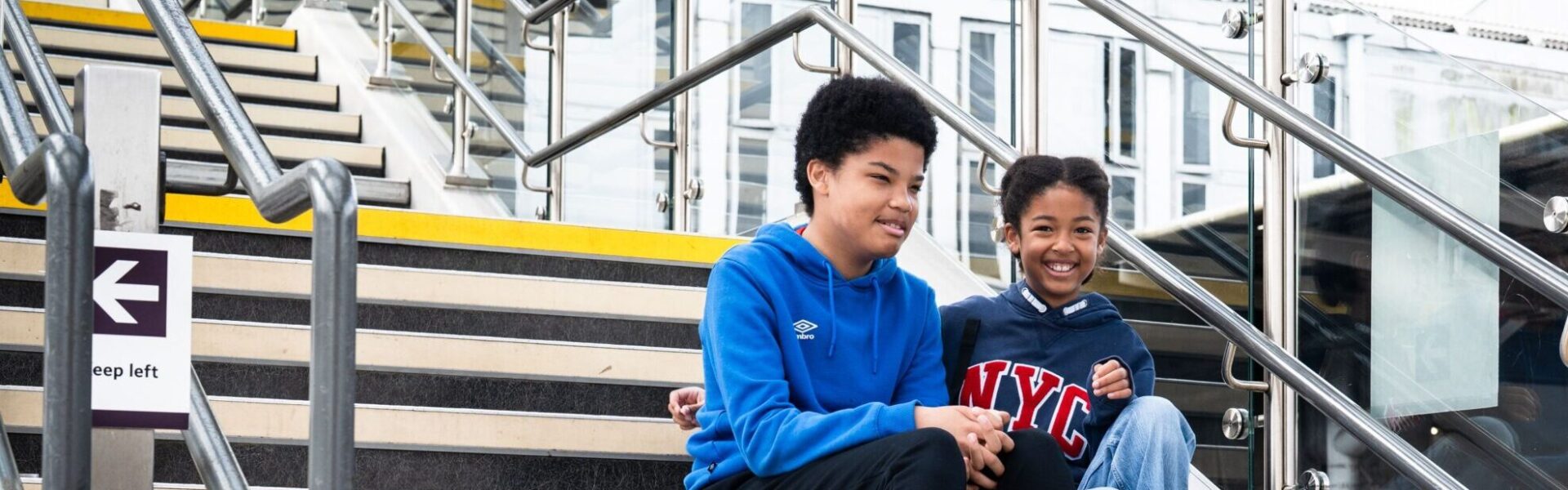Railway station steps with two young people seated together, with handrails and station structures visible around them.